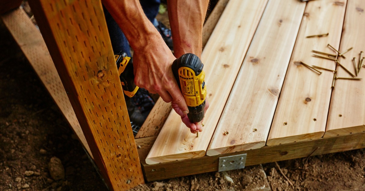 A worker measuring and drilling wood for a deck. He is using an electric drill to drive screws into the wood.