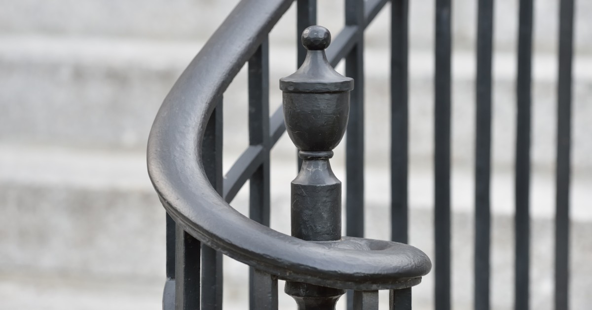 A close-up of a wrought iron staircase handrail and finial shows that the detailed finial features several different shapes.