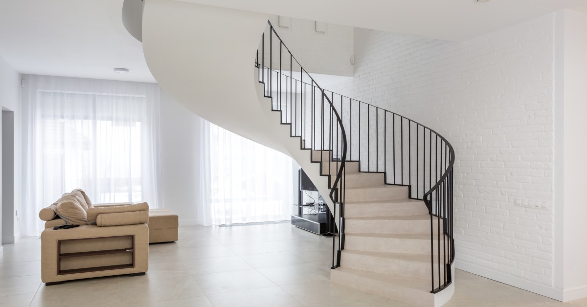 A white spiral staircase with a dark railing inside a brightly lit home. There is a beige sofa to the left of the staircase.