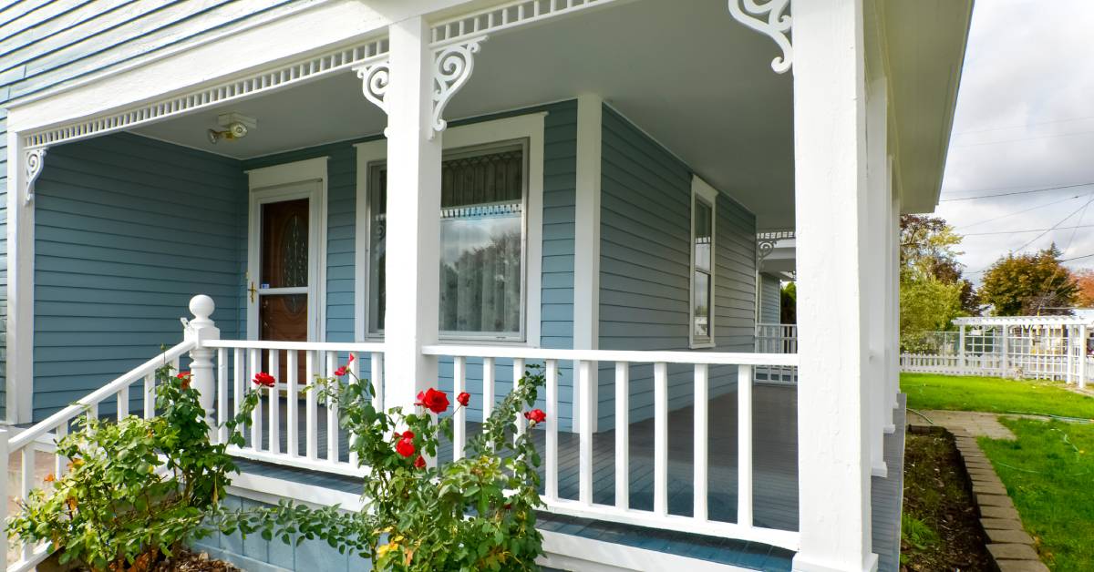 A covered wraparound porch with rose bushes in the front. The porch's railing and decorative elements are painted white.