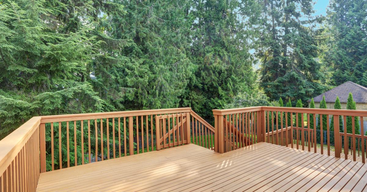 An empty wood deck with simple wood railings and a staircase. The deck is surrounded by mature green trees.