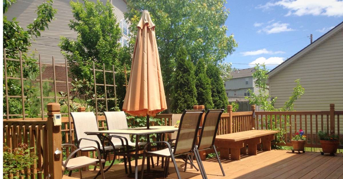 An outdoor wood deck with a table, chairs, and umbrella. The deck is surrounded by a wood fence and greenery.