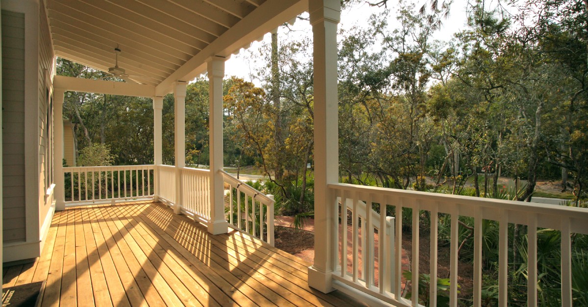 A covered wood porch with a white railing, white spindles, and white columns. The porch is overlooking a wooded area.