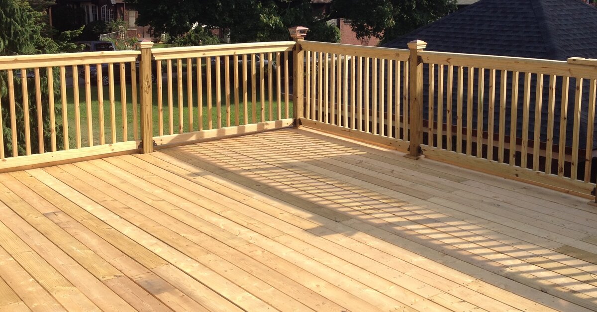 A large wooden deck surrounded by wooden railing. There are several houses and mature trees behind the deck.