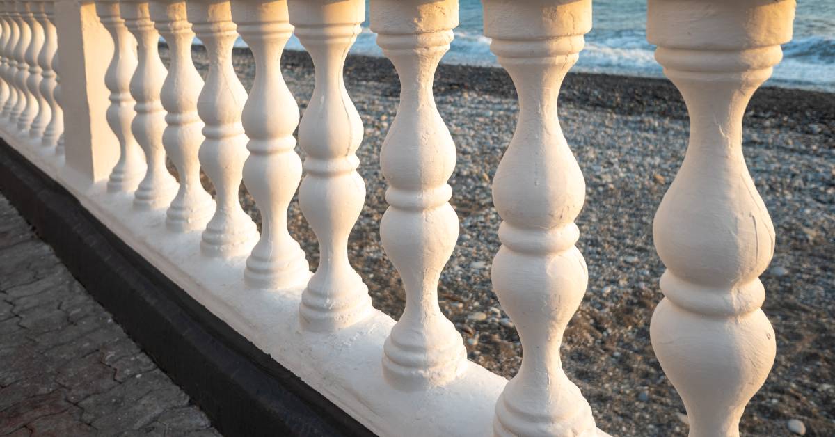 A close-up of a white railing with ornate balusters made of stone. The railing is next to a rocky beach and a body of water.