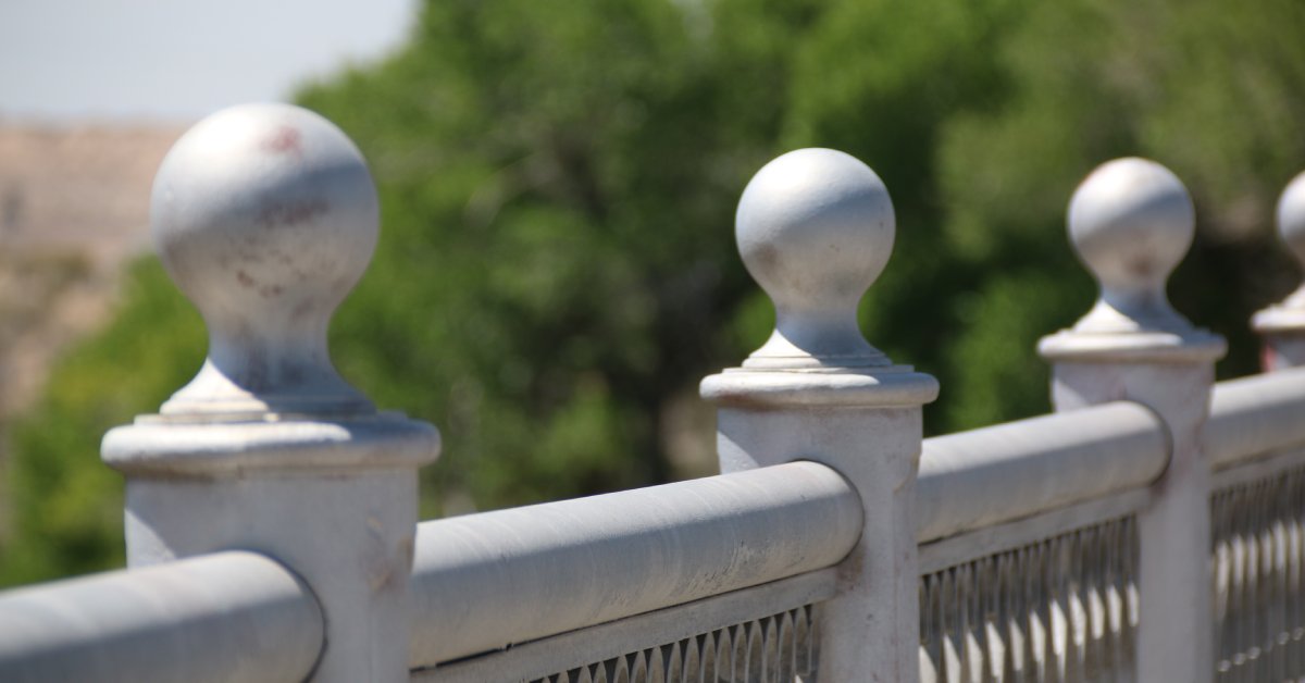 A close-up of four ball top finials sitting on the posts of a fence. There are trees behind the fence.