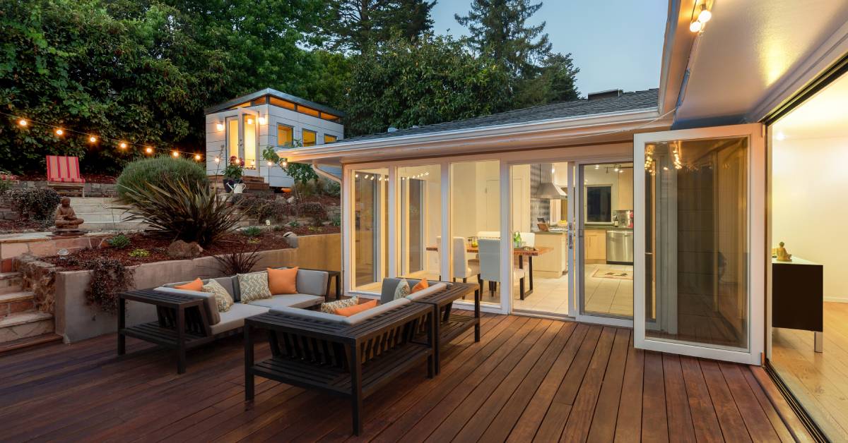 A modern home with large glass windows and doors, leading to a wooden deck with outdoor seating and multi-level landscaping.