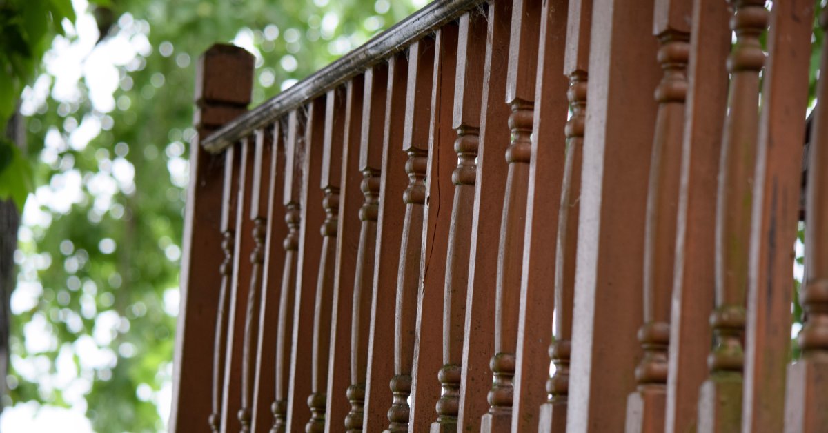 Dark stained wooden porch railings and balusters that are showing some wear and tear, with trees in the background.