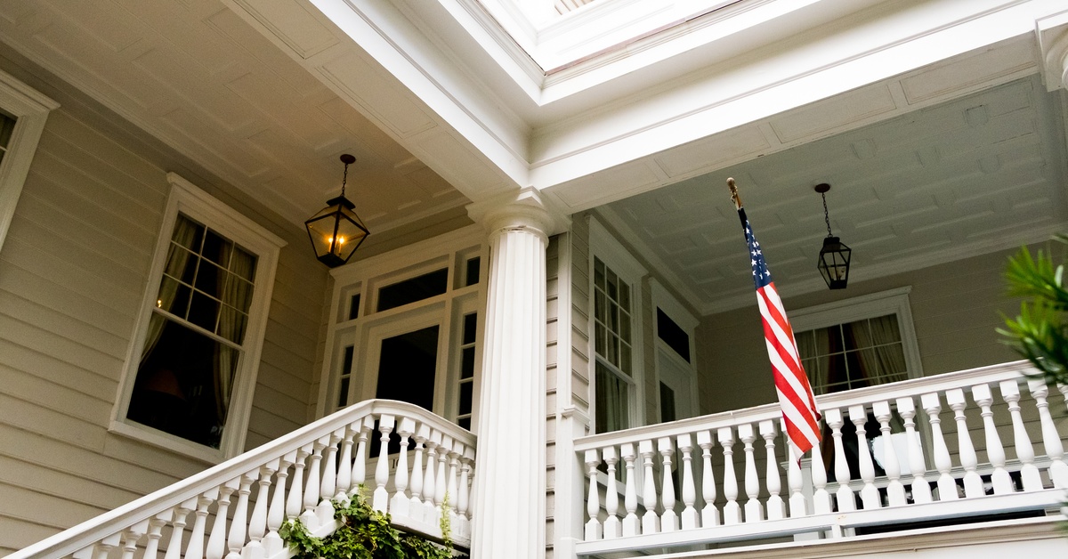 A traditional home features stairs leading to a large porch with many windows and an American flag hanging from the side.
