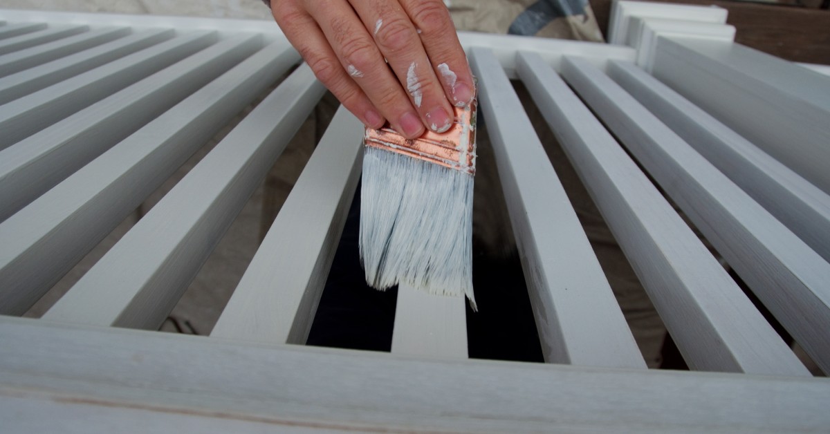 A person paints porch columns and balusters white with a brush, holding it in a hand that also has white paint on it.