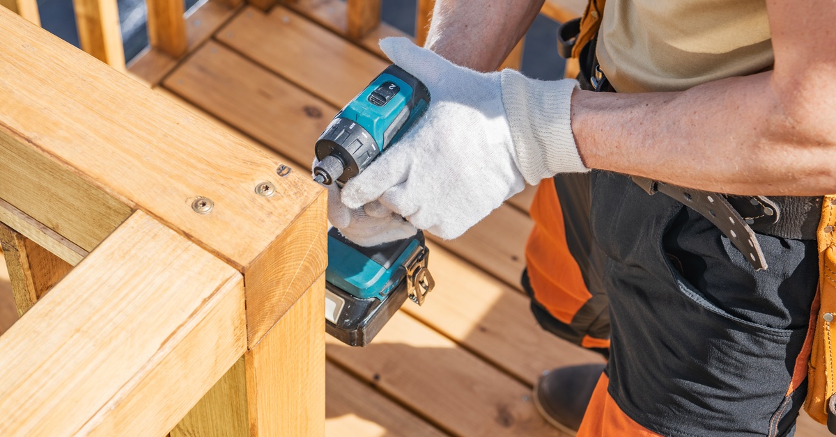 A man wearing gloves uses a drill to securely fasten a wooden deck railing while working outside on a bright, sunny day.