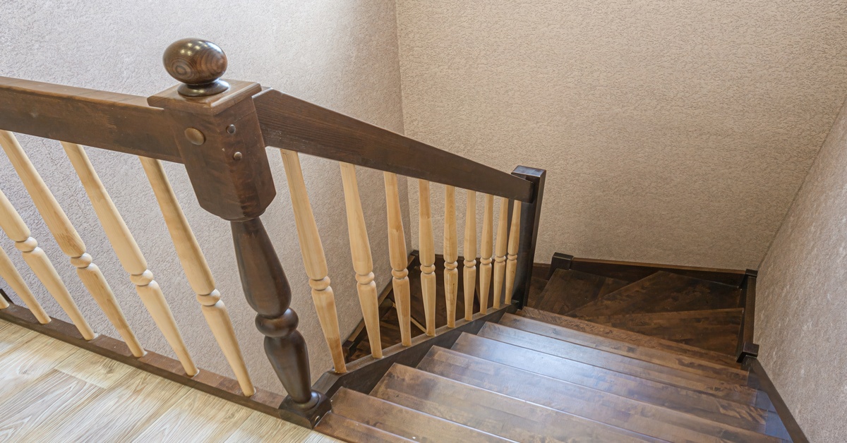 A staircase features dark and light wood stains on the railing and spindles, with wood floors and beige walls surrounding it.