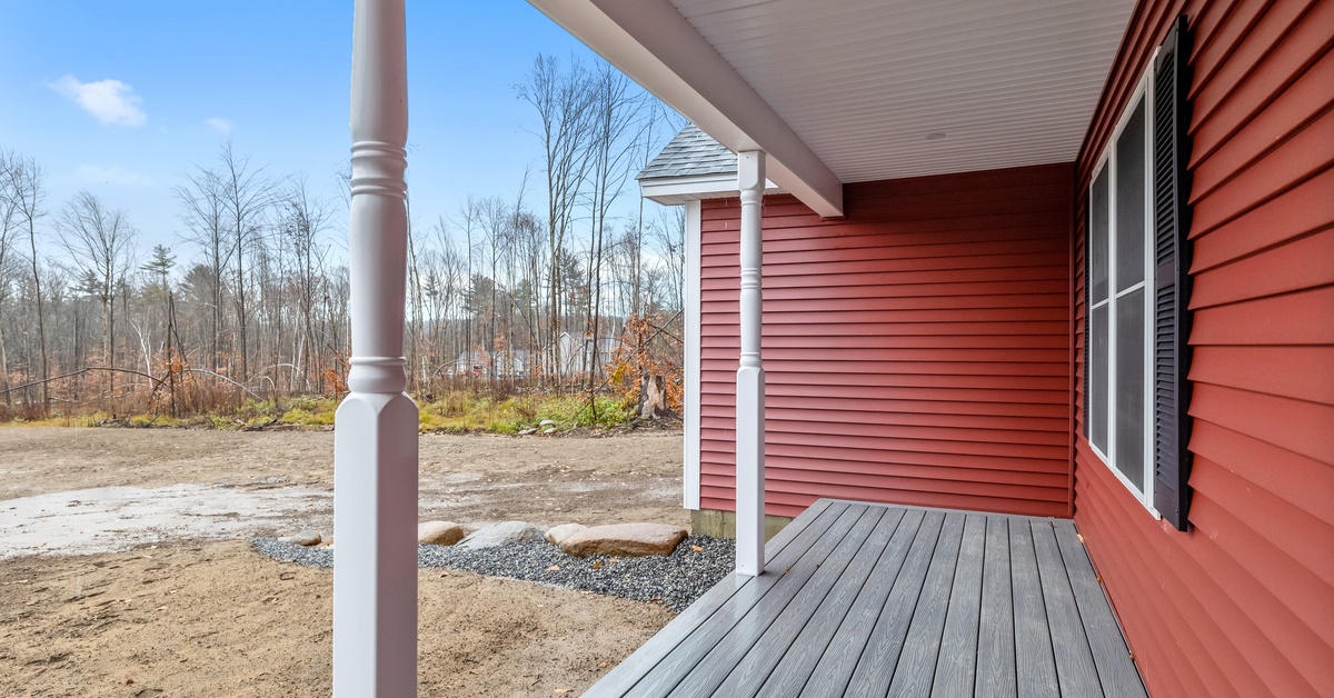 A dark wood porch with white turned posts attached to a red-sided house in late winter, with melting snow and brown grass.