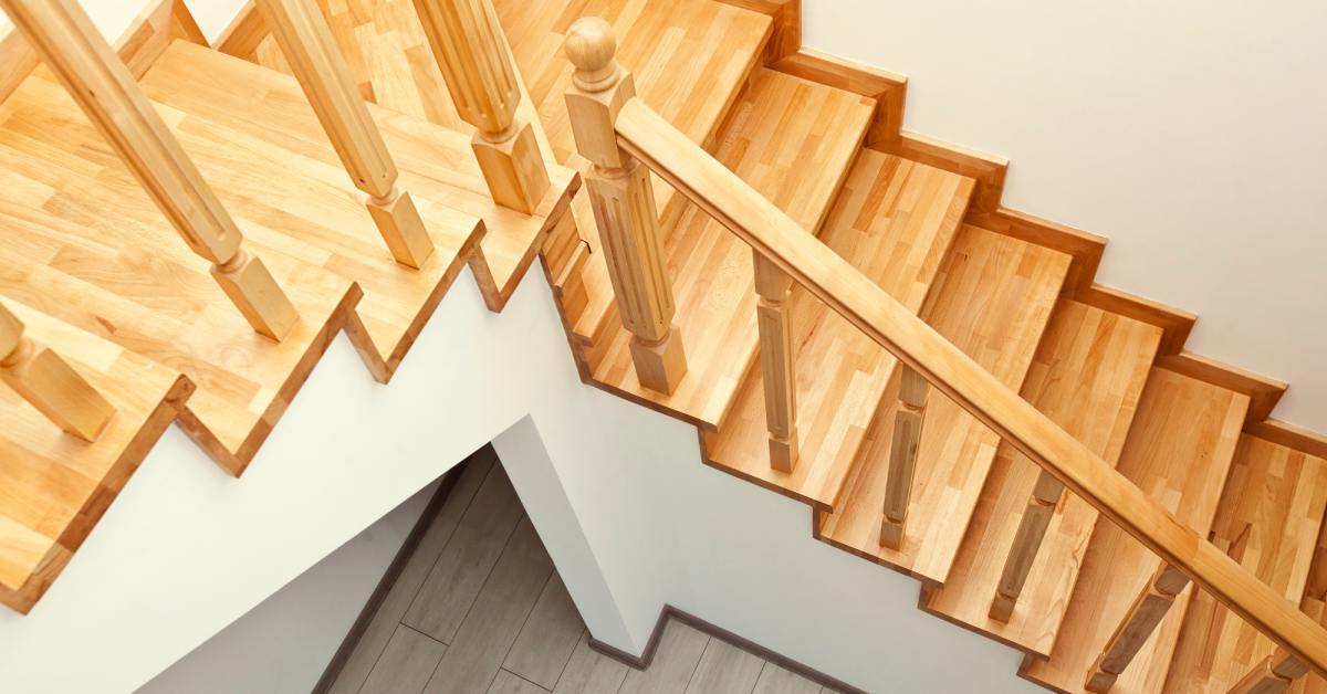 Light wood grain stairs with two flights and a platform are set against white walls and grey wood flooring below.