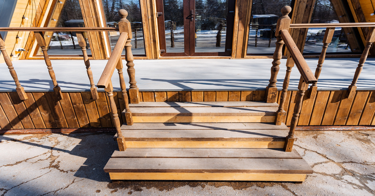 A small natural wood staircase with spindles and finial balls leads to a wooden porch of an A-frame house with many windows.