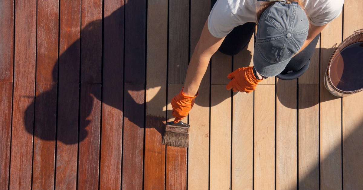 A man in orange gloves and a cap stains a light wood deck a darker color with a brush, with a can of stain beside him.
