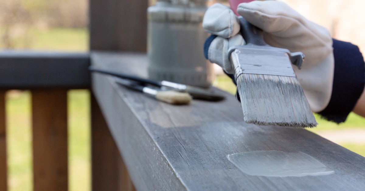 A person paints a home’s wooden porch railing gray while wearing a glove, with brushes and a paint container resting nearby.