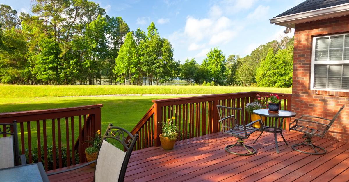 A wooden backyard deck with an outdoor table, chairs, and potted plants overlooks a grassy yard and trees.