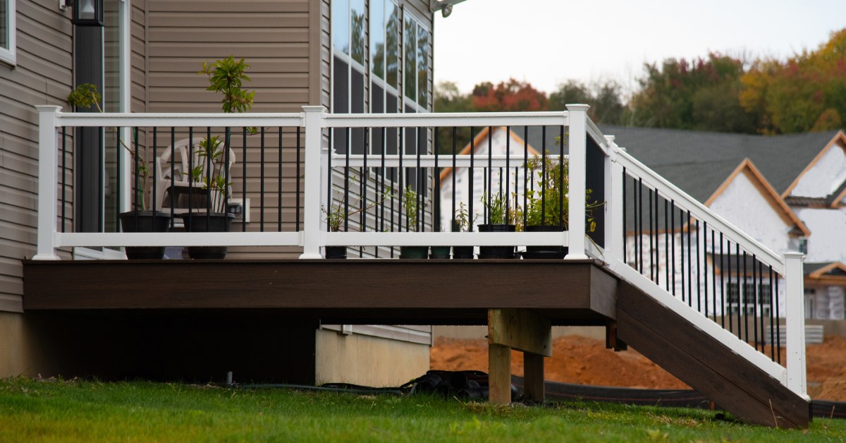A backyard deck with white railings and stairs extends from a house, with potted plants and nearby homes.