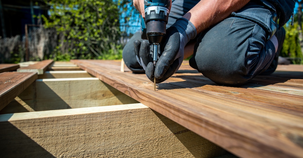 A worker using a power drill to fasten wooden deck boards, installing screws on outdoor construction framing.