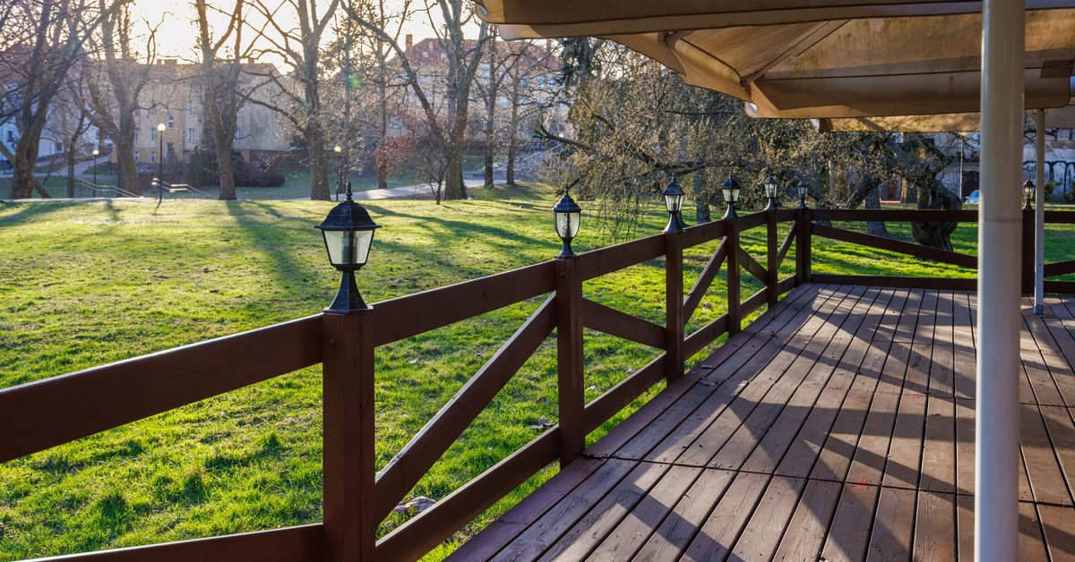 Wooden terrace with railing and lantern-style lights overlooking a sunlit park with trees, grass, and buildings.