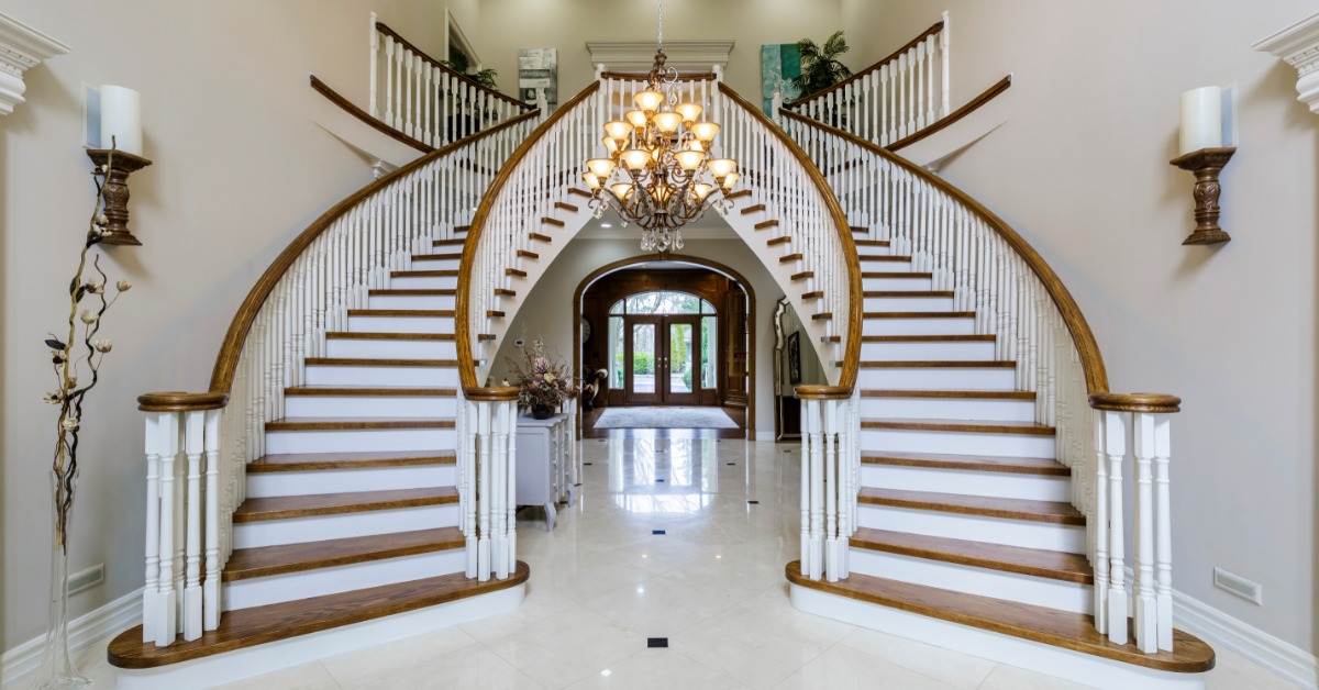 Grand foyer featuring a sweeping double staircase with white balusters, polished wood railings, and an elegant chandelier hanging above a marble floor.