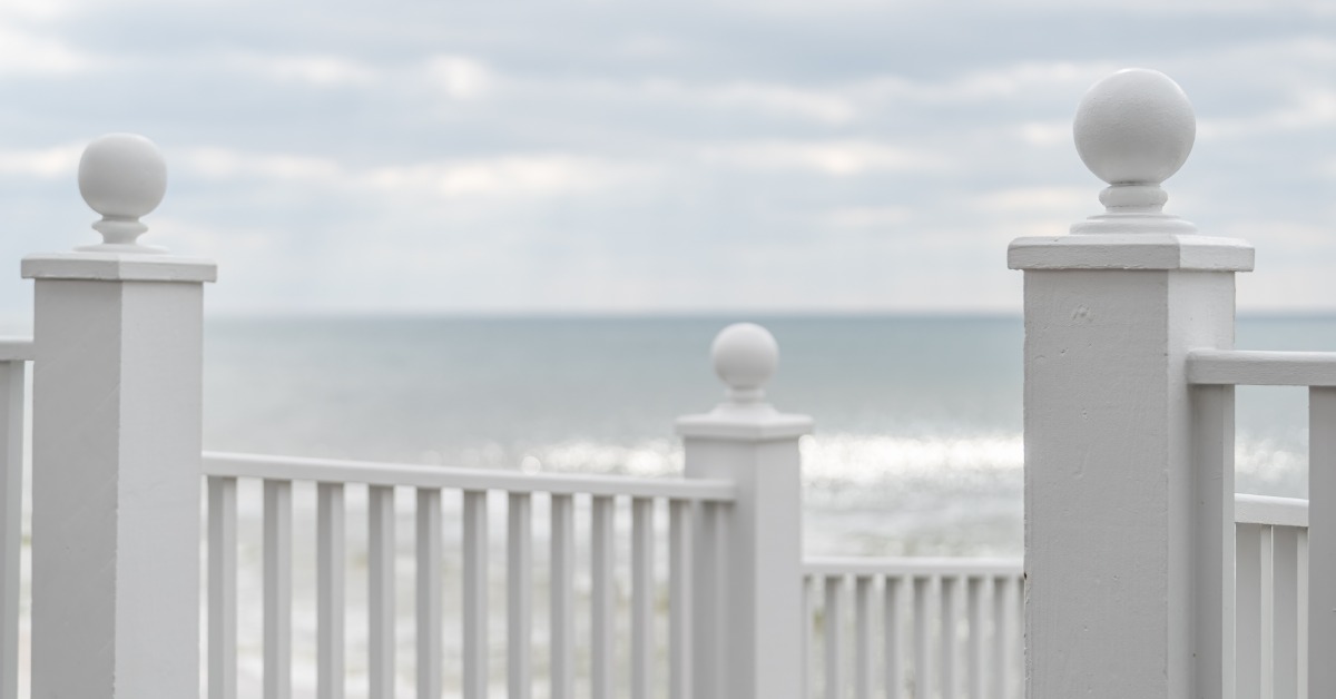 White wooden railing posts line a seaside porch overlooking calm ocean waves under a cloudy sky near the shoreline.
