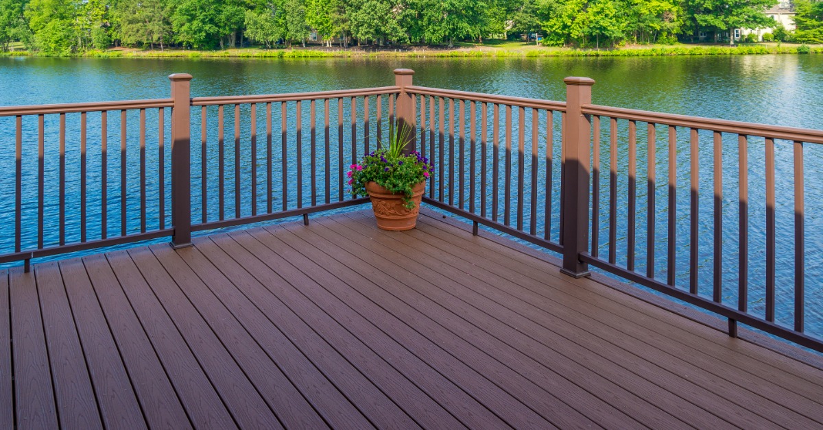 A lakeside deck features sturdy railing posts and balusters overlooking calm water bordered by mature green trees.