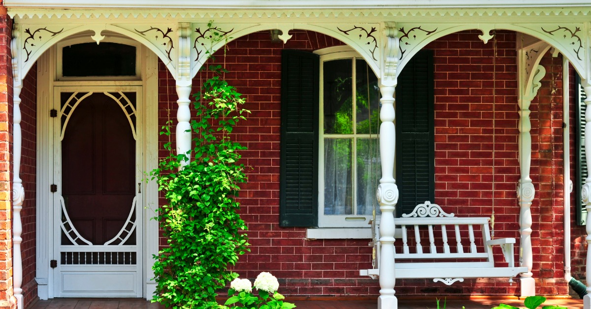 A historic brick porch features ornate white wooden trim, turned columns, and a hanging swing beneath decorative arches.